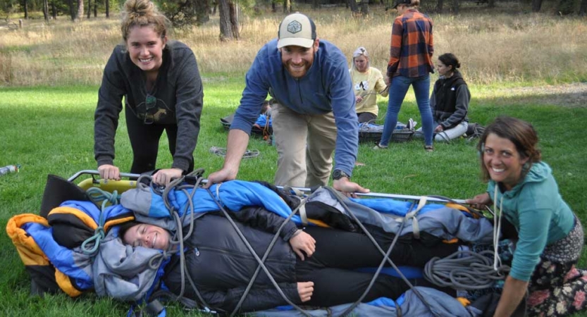 a person lies in a stretcher during a wilderness first responder training while three others smile at the camera and embrace the stretcher.